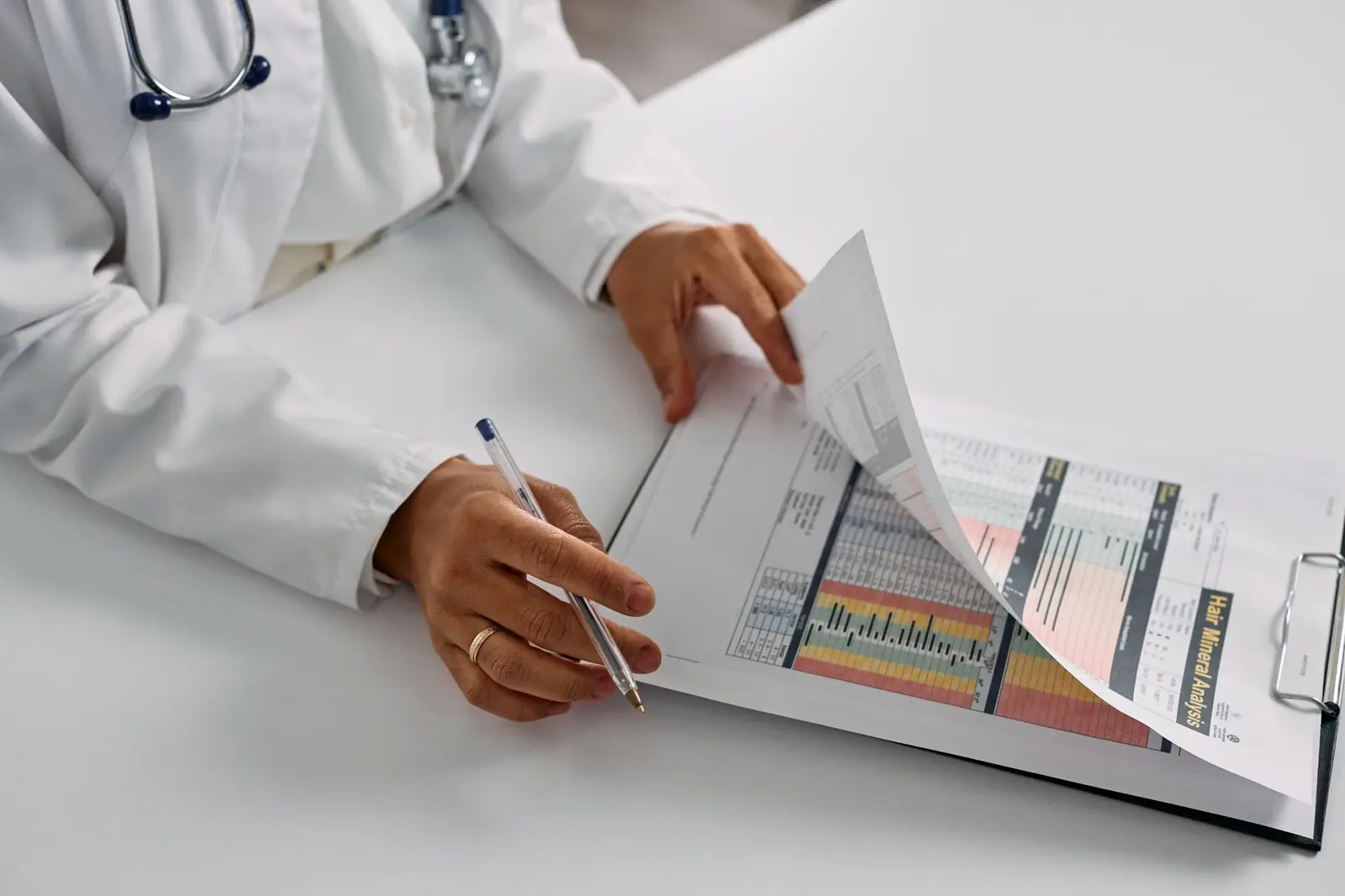 A doctor in a white coat, pen in hand, turns the pages of a printed lab report on a clipboard.