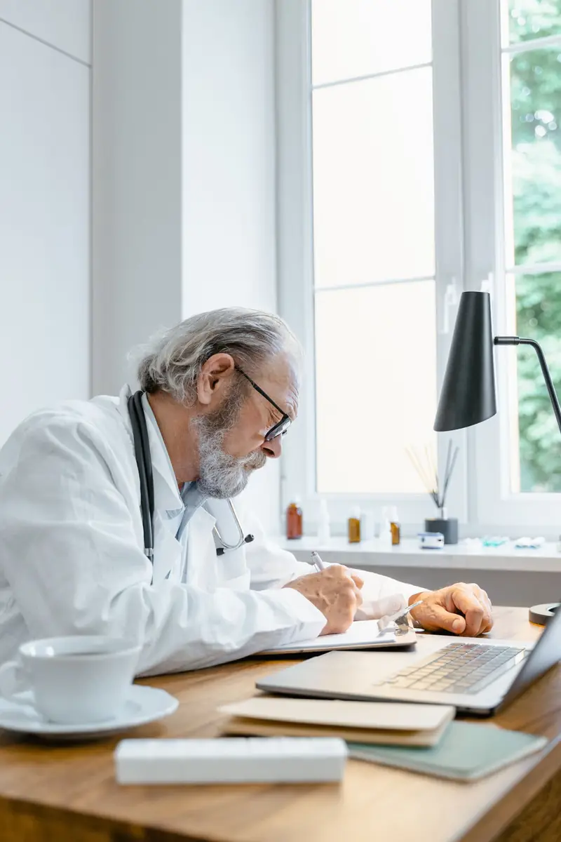 An older doctor with a stethoscope writes at a desk by a bright window, a laptop and a cup of tea beside him.