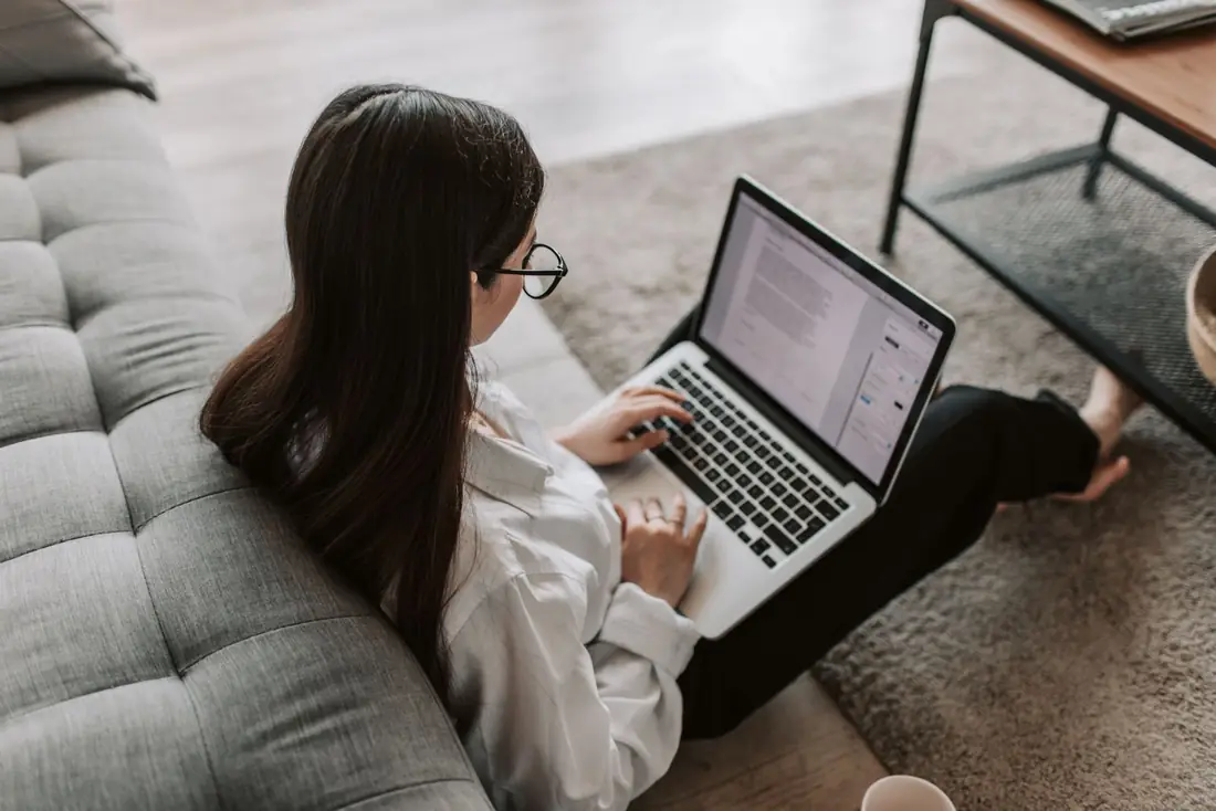 A woman sitting on the floor against a sofa, reading a document on her laptop.