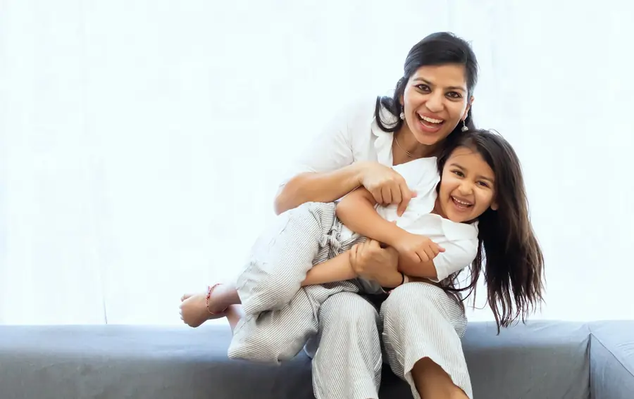 A mother and her young daughter laughing together on a grey sofa.