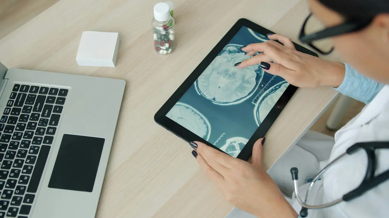 Hands swipe through brain scan images on a tablet, next to a laptop on a desk.